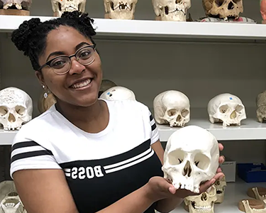 Student holding skeleton skull with rows of skulls behind her on shelves.